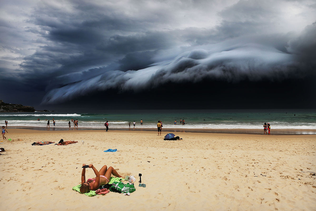 Sunbather oblivious to the ominous shelf cloud approaching - on Bondi beach. A massive “cloud tsunami” looms over Sydney in a spectacular weather event seen only a few times a year. The enormous shelf cloud rolled in from the sea, turning the sky almost black and bringing violent thunderstorms in its wake.